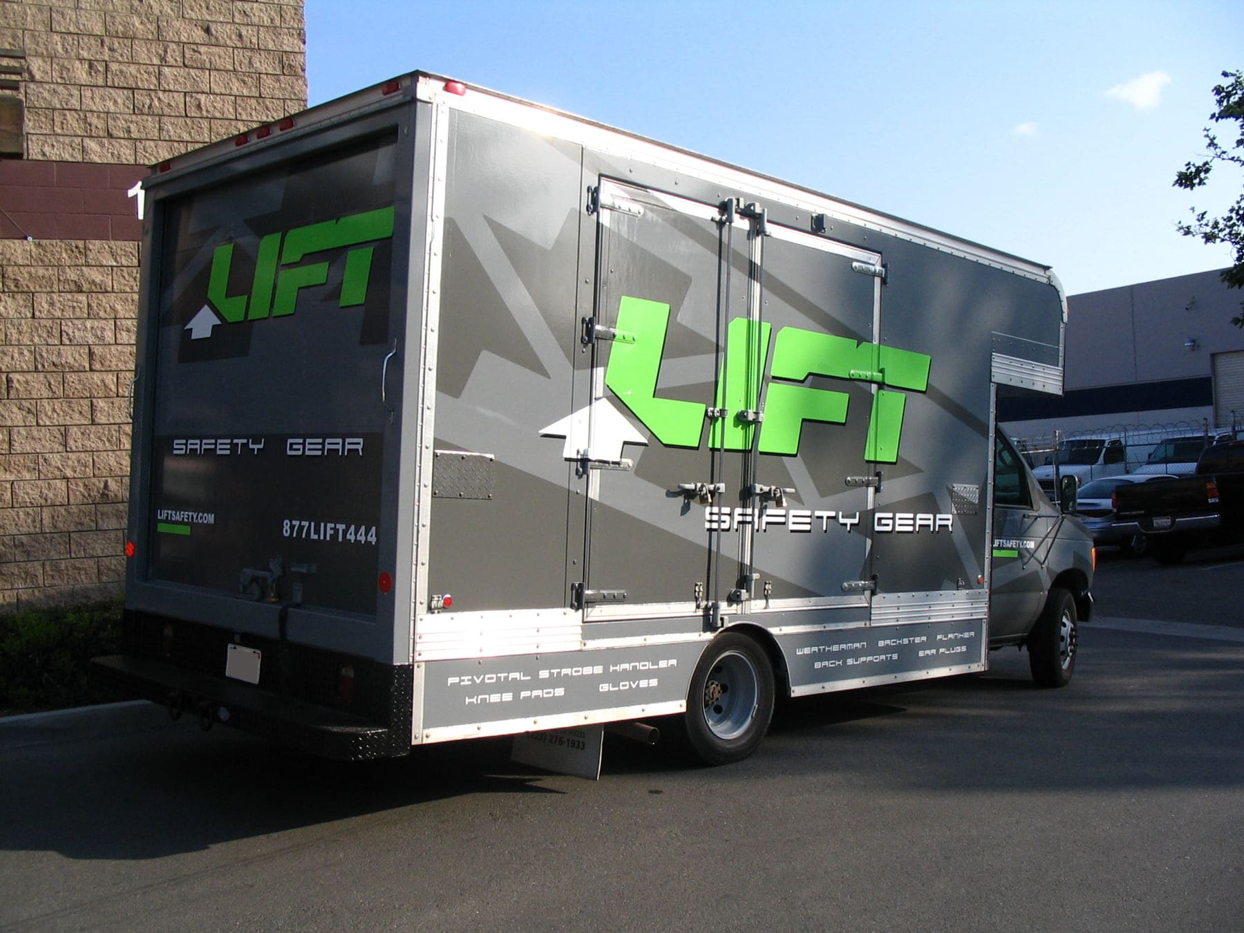 Box truck wrapped in grey and green safety-equipment graphics, parked beside a warehouse